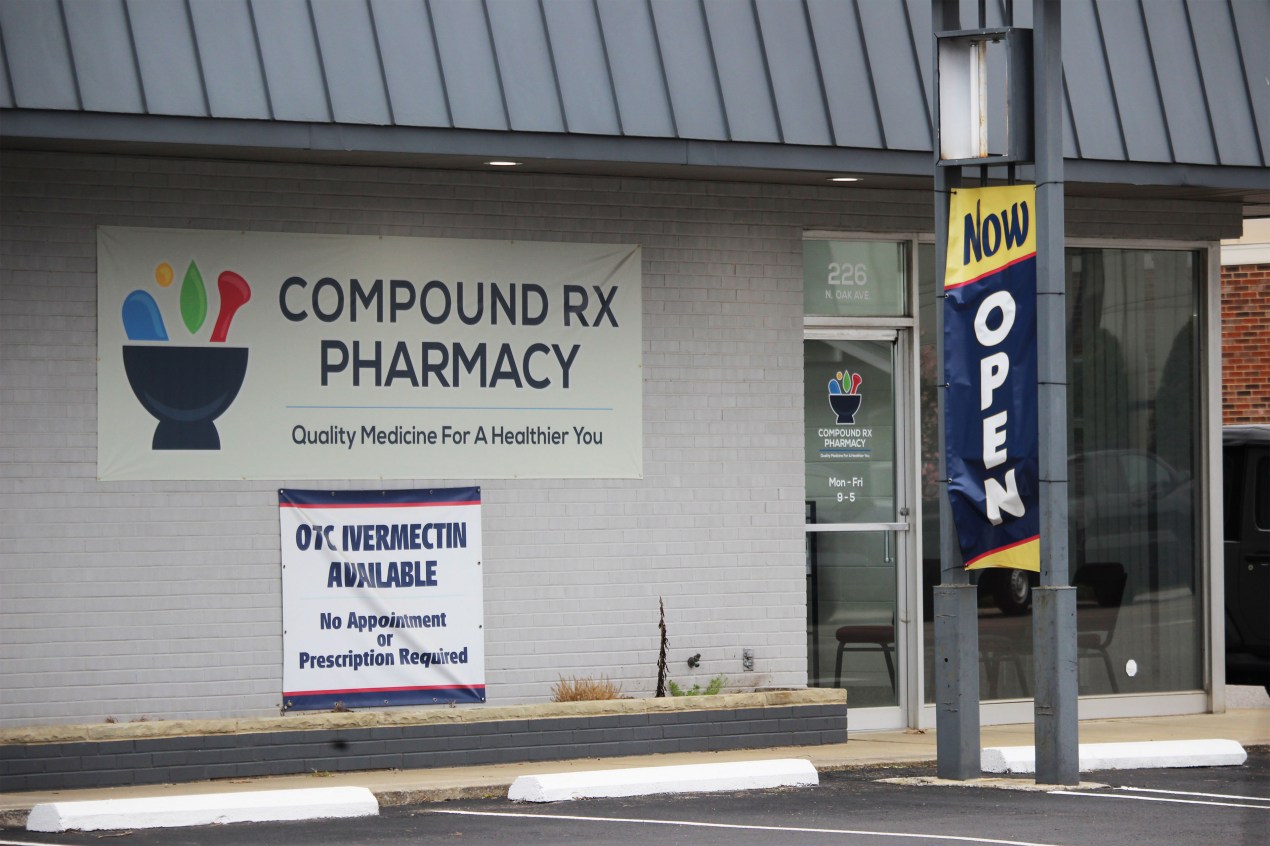 The exterior of a single-floor building. A banner on the side of it reads, "COMPOUND RX PHARMACY / OTC Ivermectin Available."