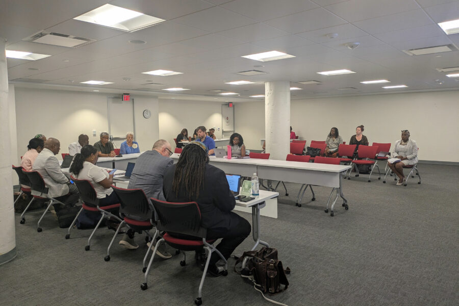 A dozen people sit around tables in a U formation in a meeting room. A few people sit in the two rows of chairs off to the side of the tables.