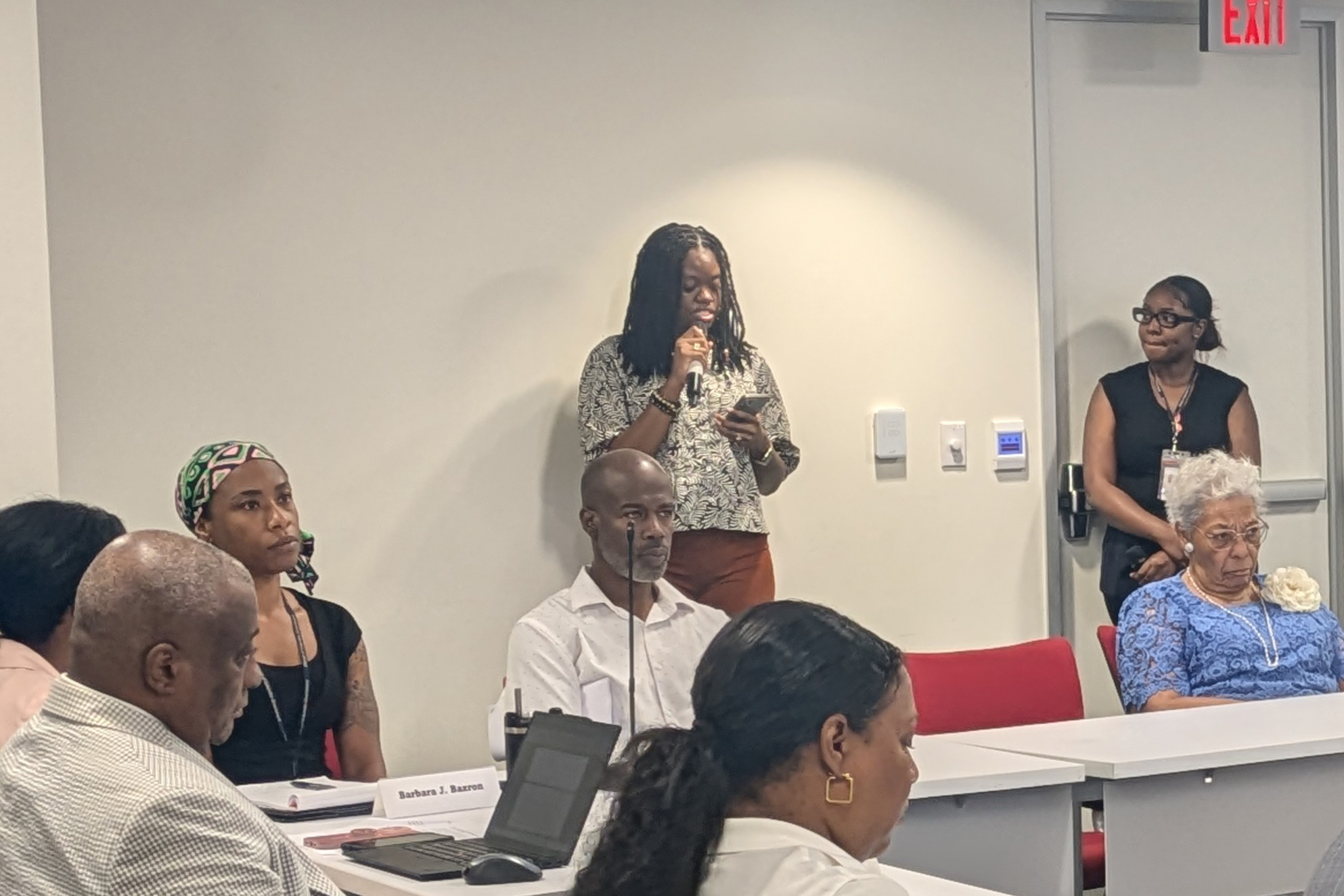 A woman speaks into a microphone. In front of her are people sitting at tables in a U formation.