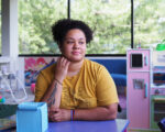 A woman wearing a yellow shirt sits at a blue table looking out toward a window