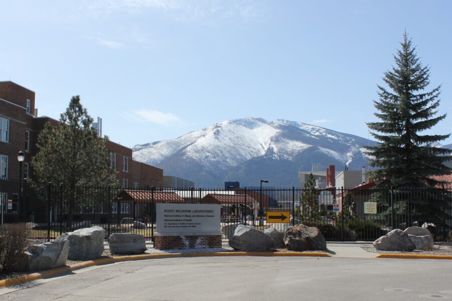 The metal gate with a sign that reads "Rocky Mountain Laboratories" framed by pine trees and snow capped mountain in the background