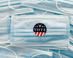 A photo of a pile of disposable surgical face masks. A sticker that reads, "Vote," is placed on the central mask.
