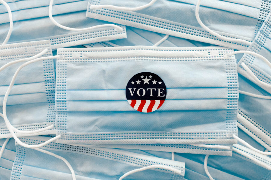 A photo of a pile of disposable surgical face masks. A sticker that reads, "Vote," is placed on the central mask.