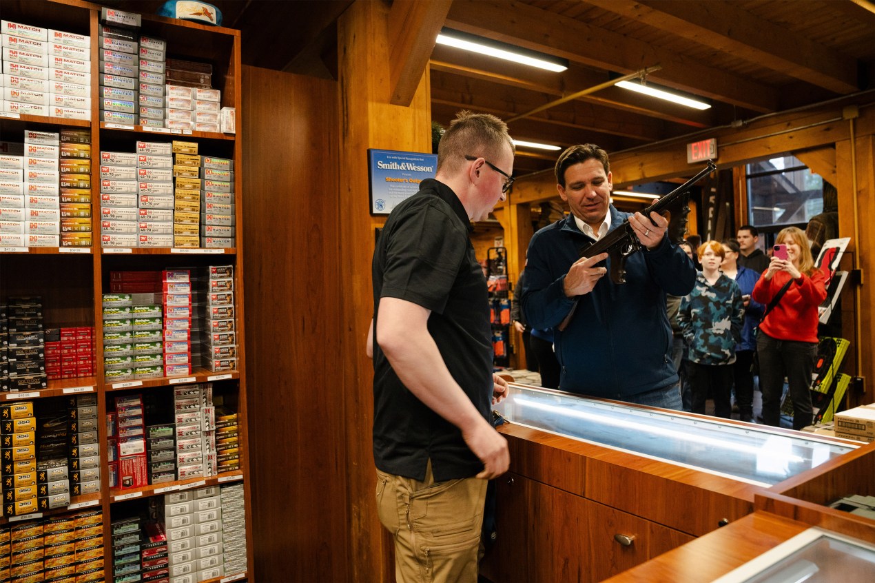 Ron DeSantis examines a Thompson submachine gun at a gun shop during his presidential run in 2023. A crowd of people watch behind him.