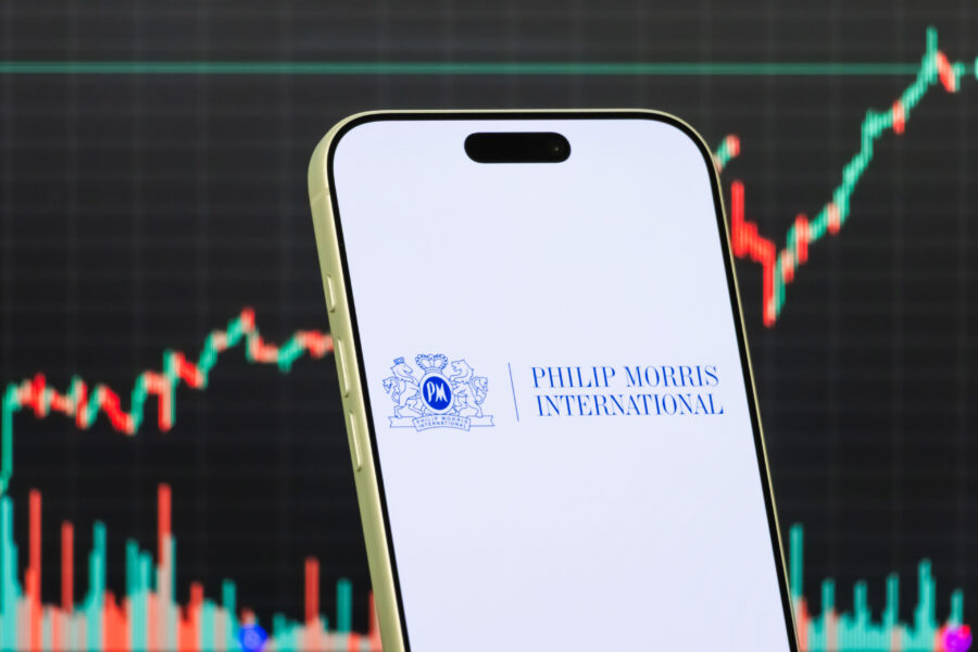 A smartphone displays the logo of Philip Morris International Inc. in front of a screen showing the company's latest stock market chart, which has increased in value.