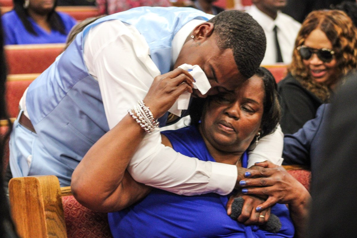 A man leans over to hug a woman who is sitting in front of him. The woman gently touches a tissue to the man's face in an emotional moment.