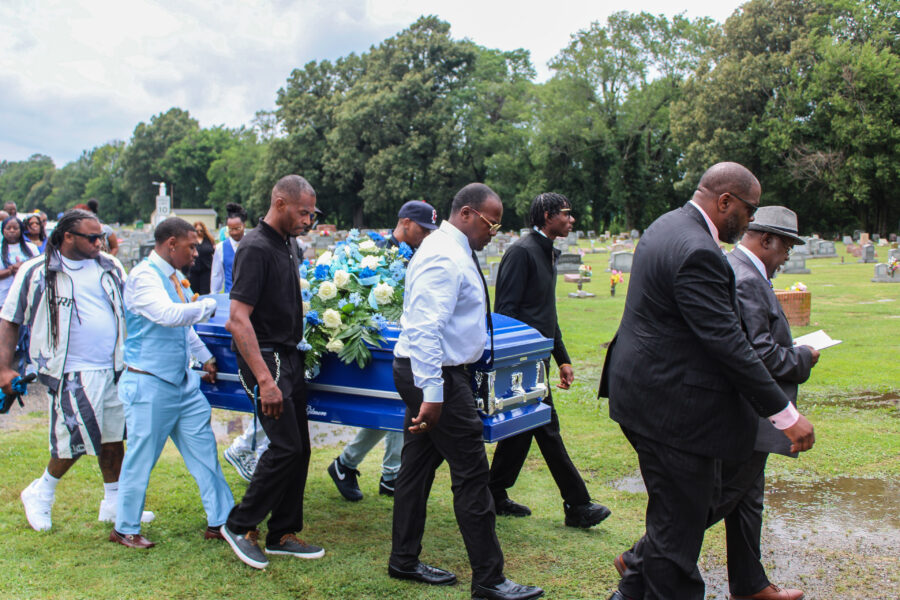 A small group of men wearing black and pale blue carry a casket.
