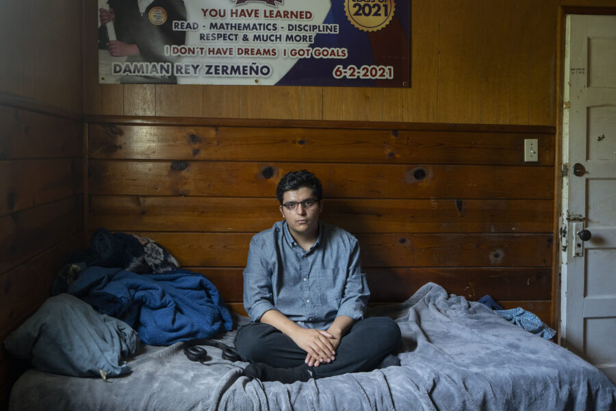 A man with dark hair and glasses sits on a bed with a blue blanket as he looks at the camera