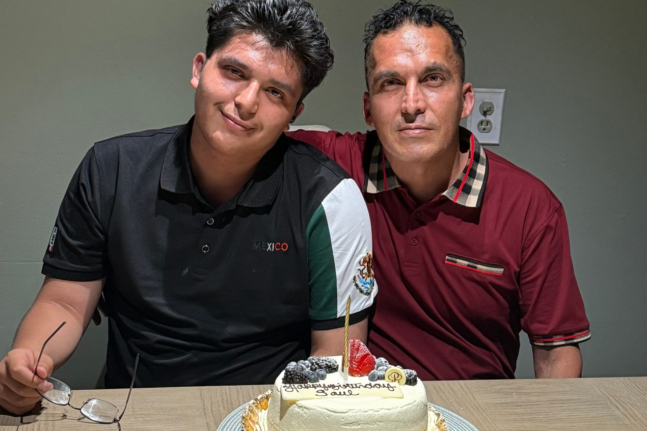 Two men sit at a table in front of a birthday cake as they smile for a portrait