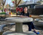 A woman wearing a dark shirt and pants, a bright blue shoes sits at a stone chess table in a park and looks at the camera. A playground is in the background.