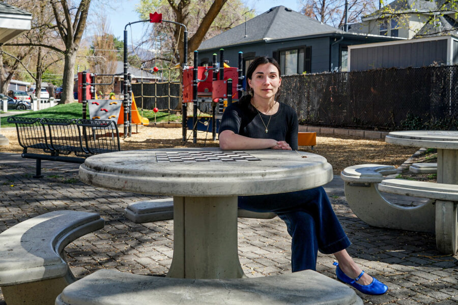 A woman wearing a dark shirt and pants, a bright blue shoes sits at a stone chess table in a park and looks at the camera. A playground is in the background.