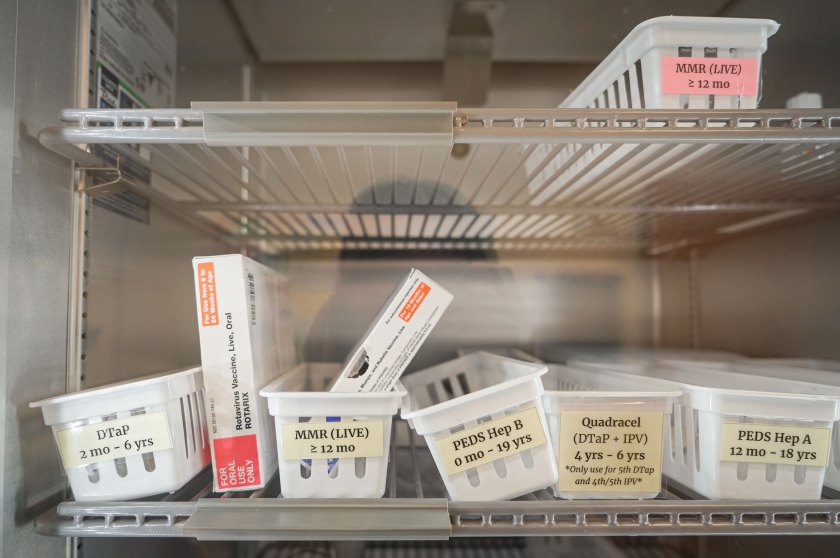 measles-doctors-06-vaccines_web A close-up image through a glass door of the inside a refrigerator where white plastic containers with labels such as "MMR (LIVE) greater than or equal to 12 mo" are lined up on shelves.