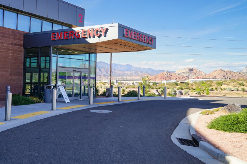 measles-doctors-07-hospital_web The emergency department entrance to a hospital is shown in the foreground and mountains are visible in the distance behind it.