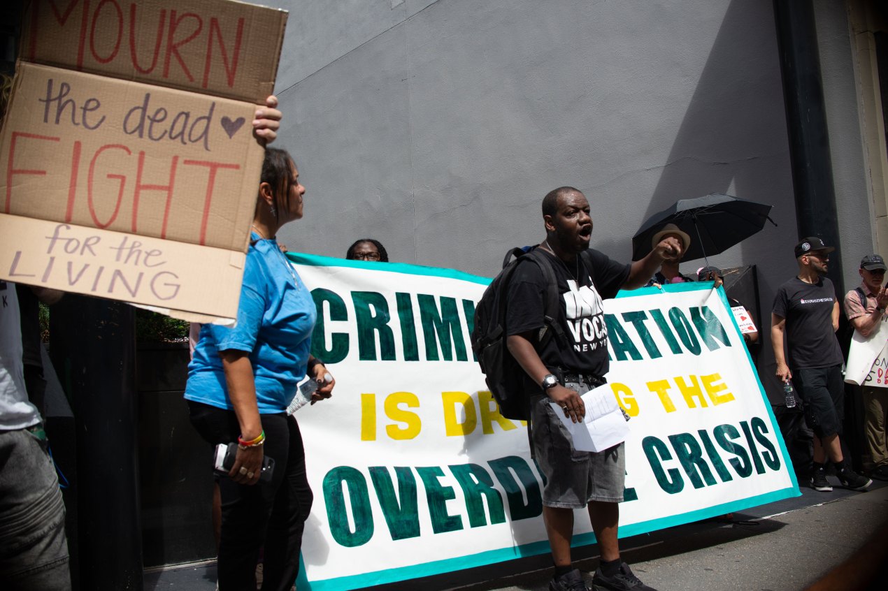 A man speaks in the middle of a group of people protesting. Behind him, several people hold a banner that is had to read because words are obscured. Another person to the left of the man holds a cardboard sign that reads, "Mourn the dead, fight for the living."