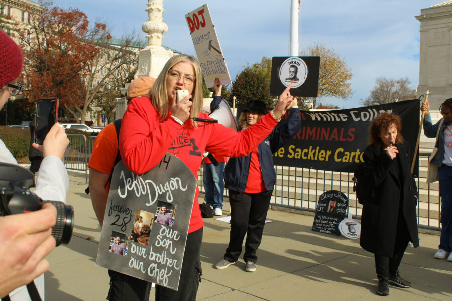 A woman at a protest holds a sign reading "Jeff Dugon, forever 28, our son, our brother, our chef" and speaks into a megaphone.
