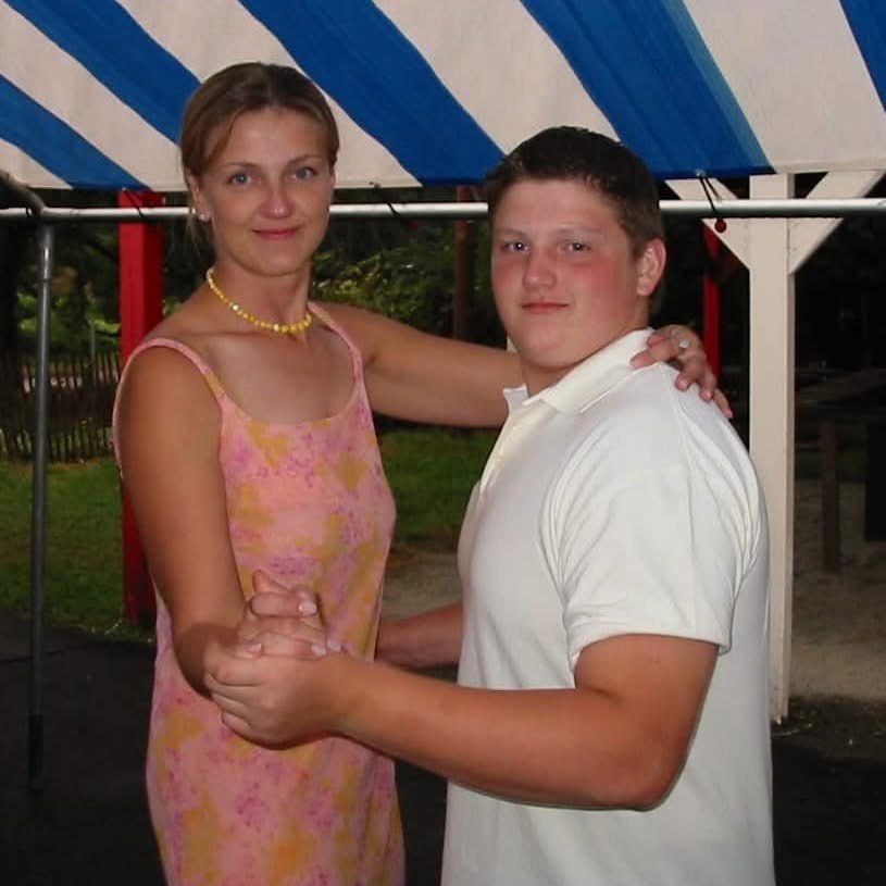 A woman in a pink dress and a teenage boy in a white polo shirt dance together under a blue and white striped awning. They both look at the camera and smile.