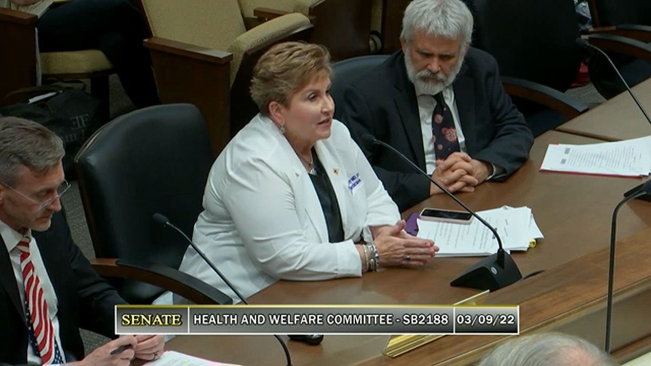 A woman wearing a white jacket sits at a courtroom table with two men in suit jackets beside her.