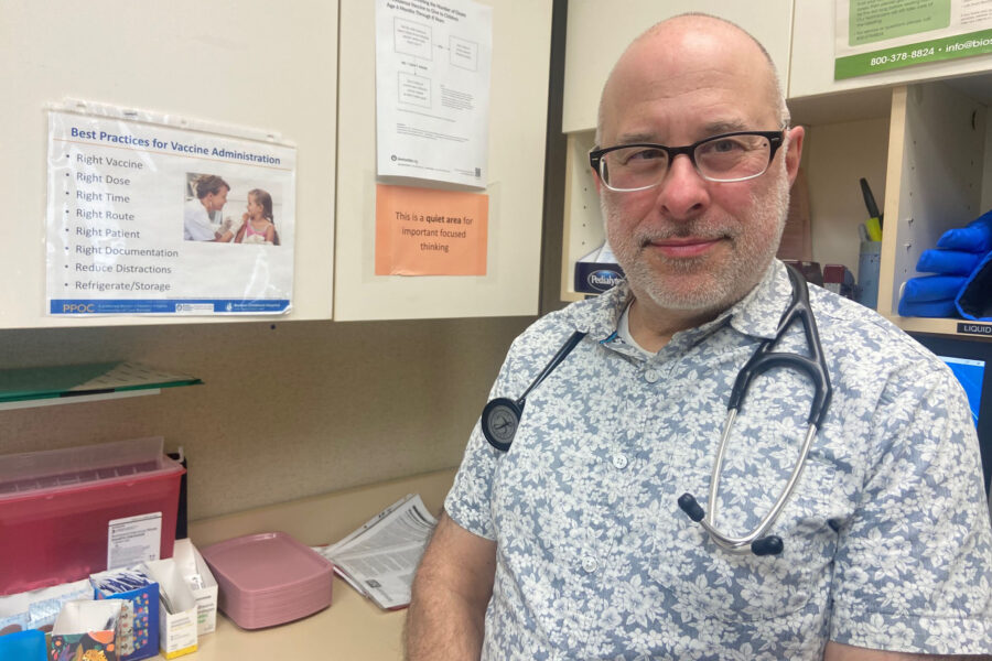 A male doctor wearing a short sleeve button down and stethoscope around his neck stands for a photo in his medical office.