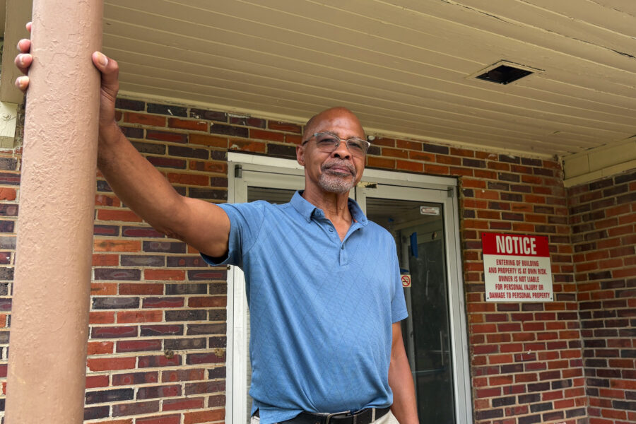 A Black man stands on a porch of a brick building.