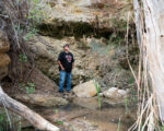 A man stands in front of a natural spring of water, surrounded by rocks and vegetation.