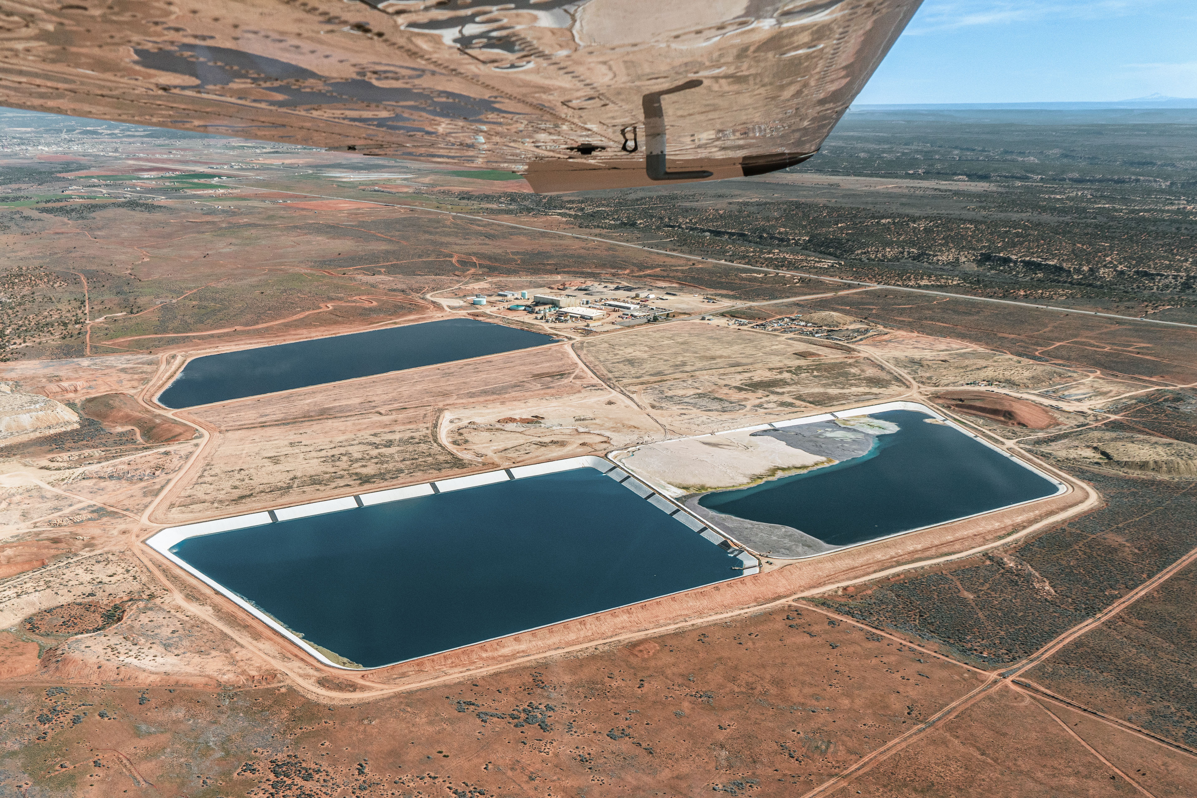 An aerial view shows  tailing ponds amidst a mesa landscape. The wing of an airplane from which the photo was taken is seen in the frame.
