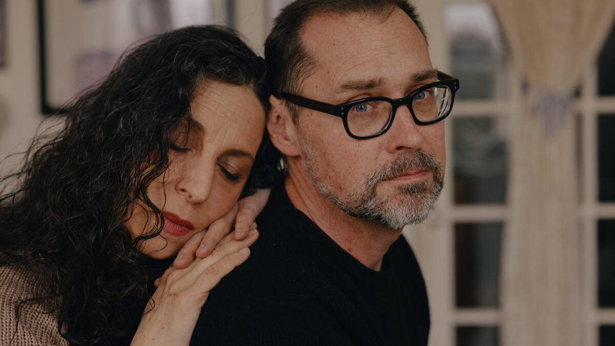 A woman with dark hair leans with her eyes closed on the back of a man wearing glasses who looks at the camera