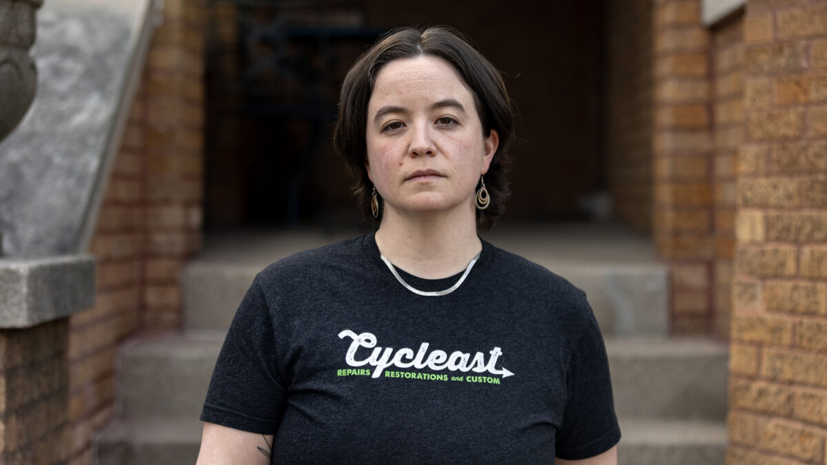 An adult with short brown hair, wearing a black T-shirt, stands in front of a stoop and is directly facing the camera.