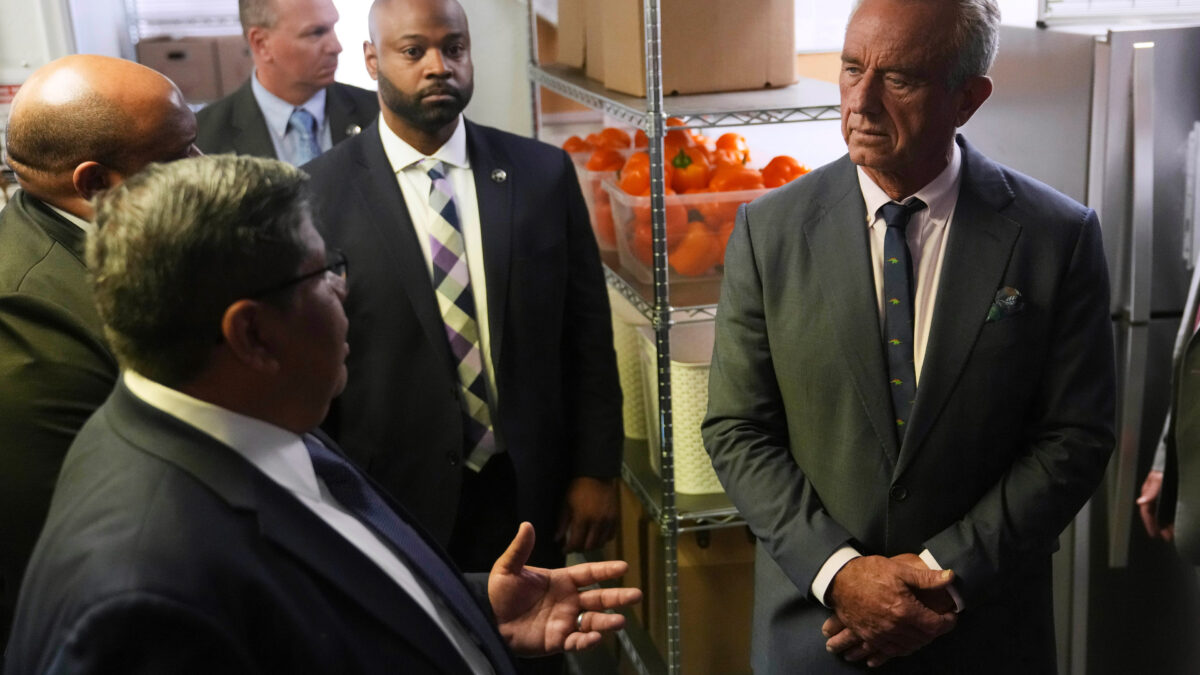 A photo of Robert F. Kennedy Jr. receiving a tour of a food distribution center. Shelves behind him show crates filled with orange bell peppers.
