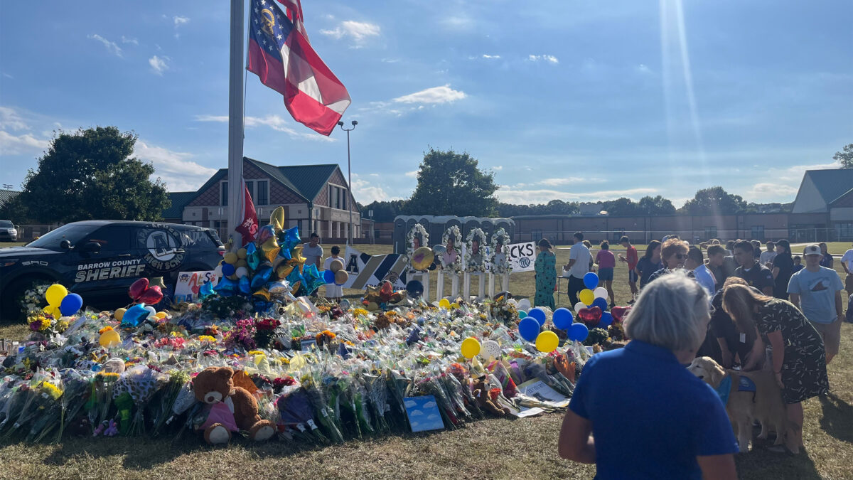 A photo of a flagpole surrounded by bouquets and stuffed animals outside of a school.