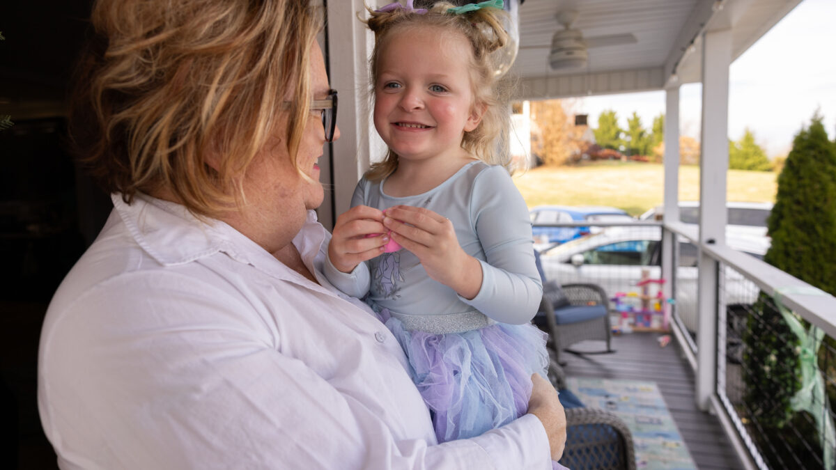 A mother holds her 3-year-old daughter in her arms on their porch. The daughter is wearing a big smile.