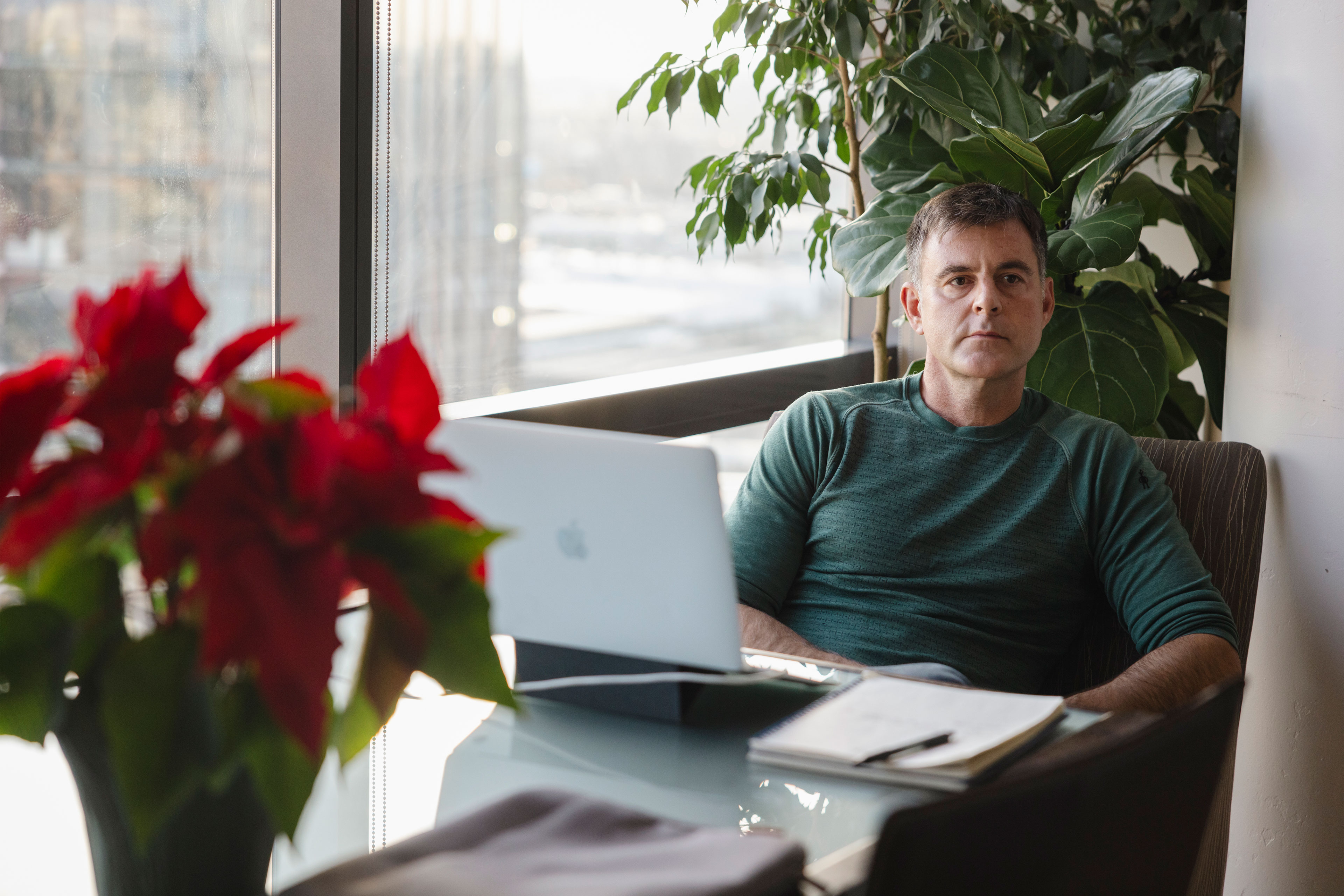 botm-dec2024-06_3840x2560web A man sits at a table near a window. A poinsettia plant, laptop, and notebook with pen rest on the table in front of him.