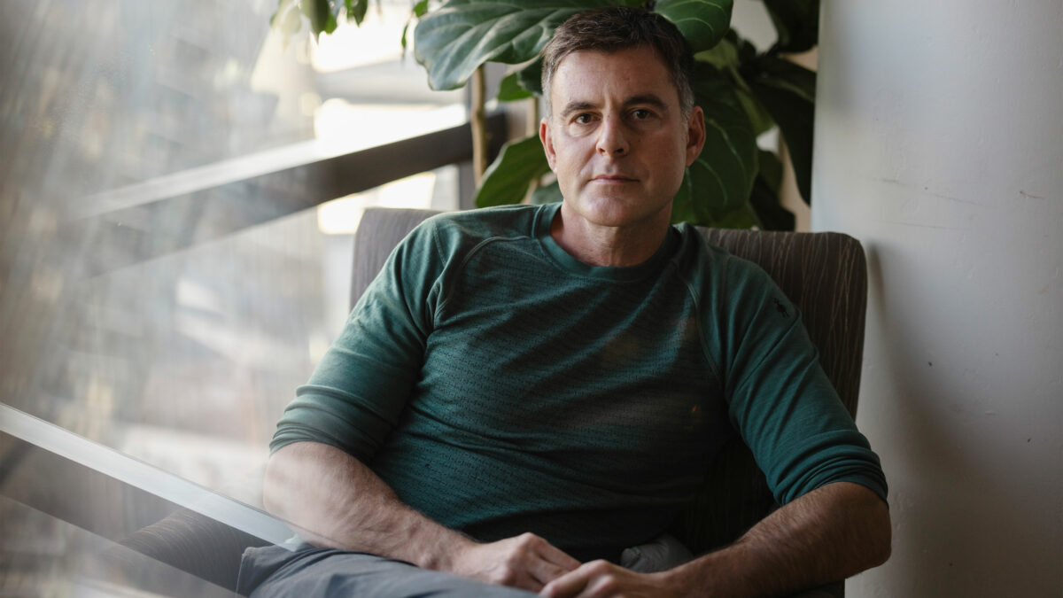A man sits in an armchair near a window and looks at the camera.