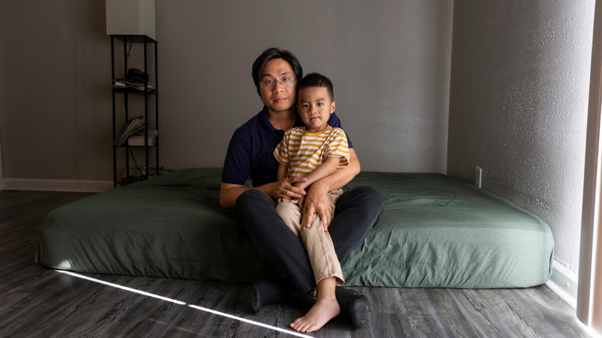 A young boy sits on his father's lap on a mattress covered in a green fitted sheet on the floor.