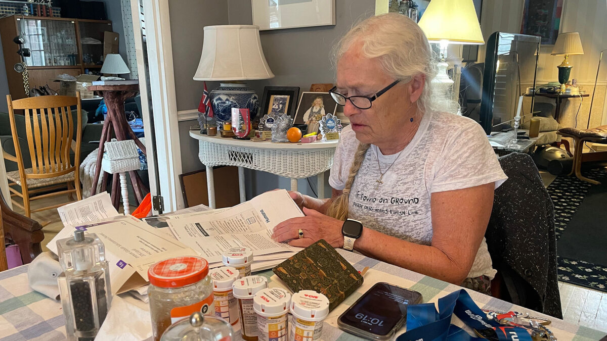 A woman with white hair in a braid sits at a dining room table and looks through paperwork. A notebook, pill bottles, a phone, and a medal on a ribbon are on the table in front of her.
