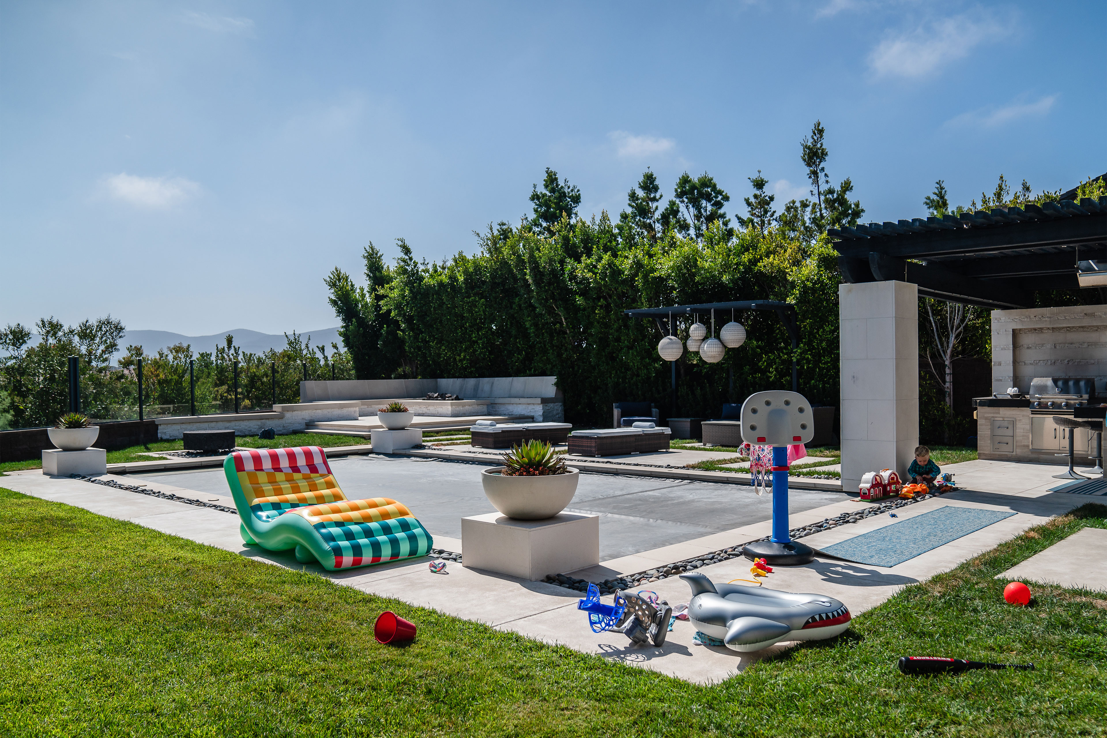 botm-oct2024-01_3840x2560web A fenced-in backyard shows a covered pool, firepit with seating, a pool chair, and toys.