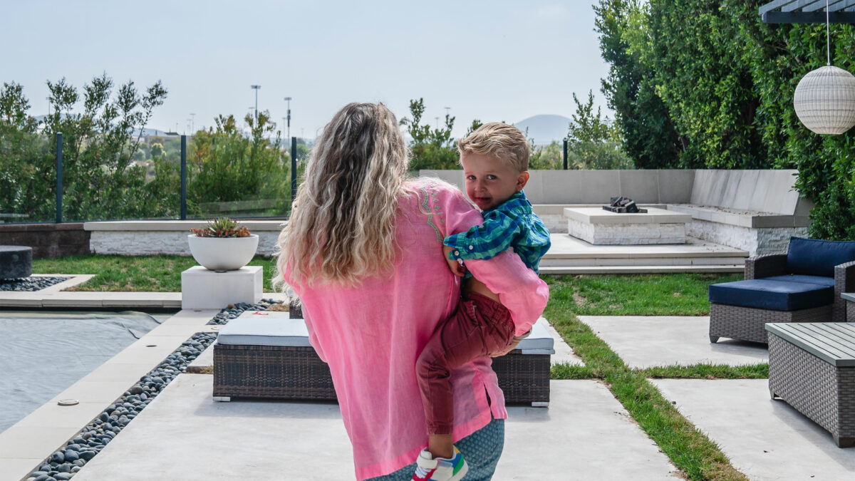 A woman in a pink shirt walks away from the camera while holding a young boy on her right him. The boy looks behind her at the camera.