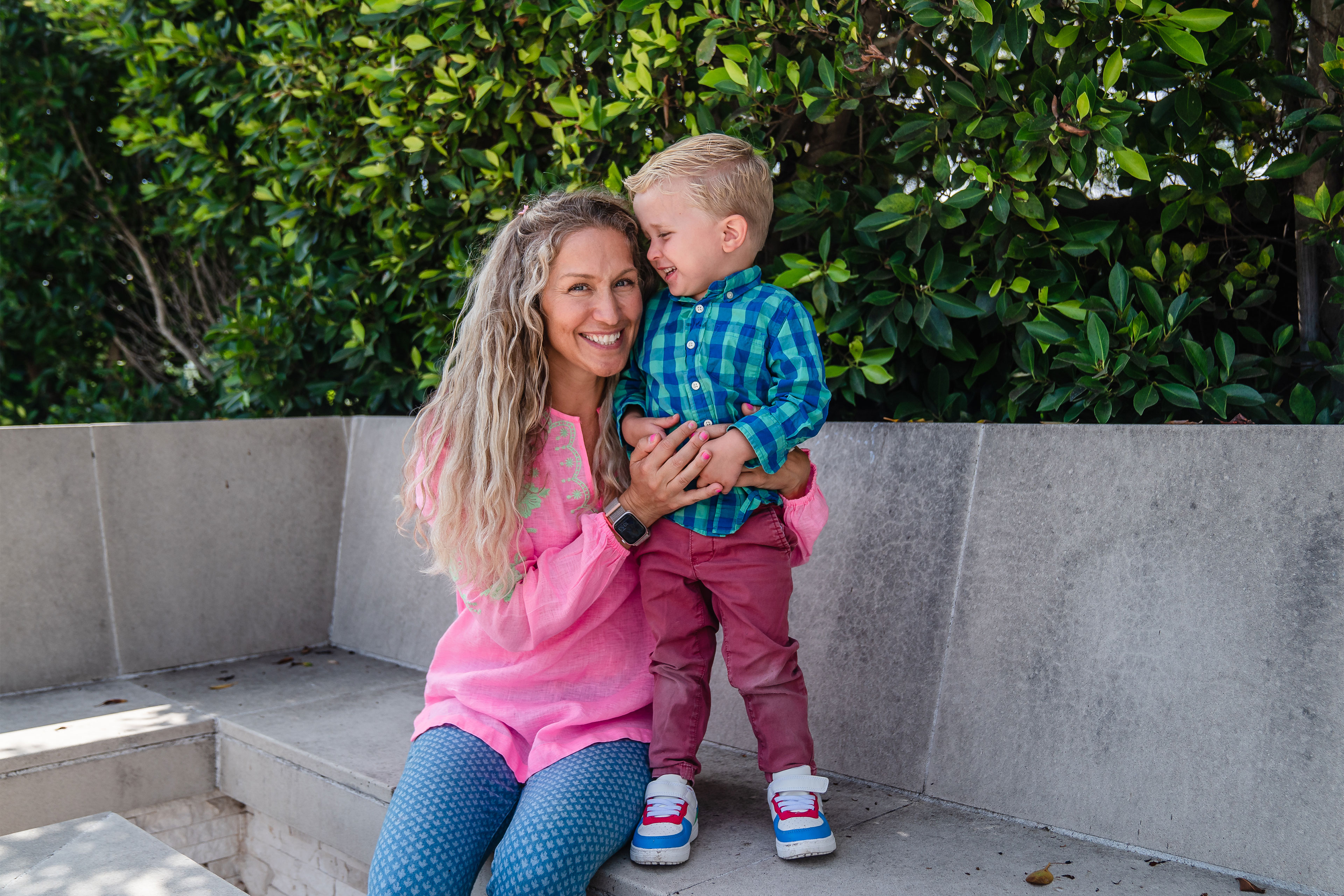 botm-oct2024-13_3840x2560web A woman sits on a concrete bench with her arm around a young boy standing on the bench beside her. She smiles at the camera and he smiles at her.