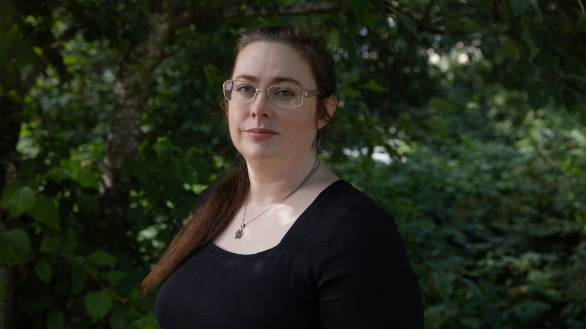 A photo of a woman standing outside with green foliage behind her.