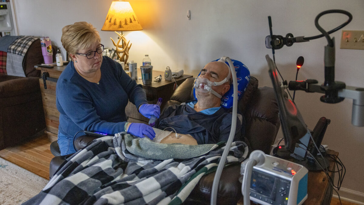 A woman wearing blue nitrile gloves helps a man who is sitting in a recliner next to her wearing a ventilator
