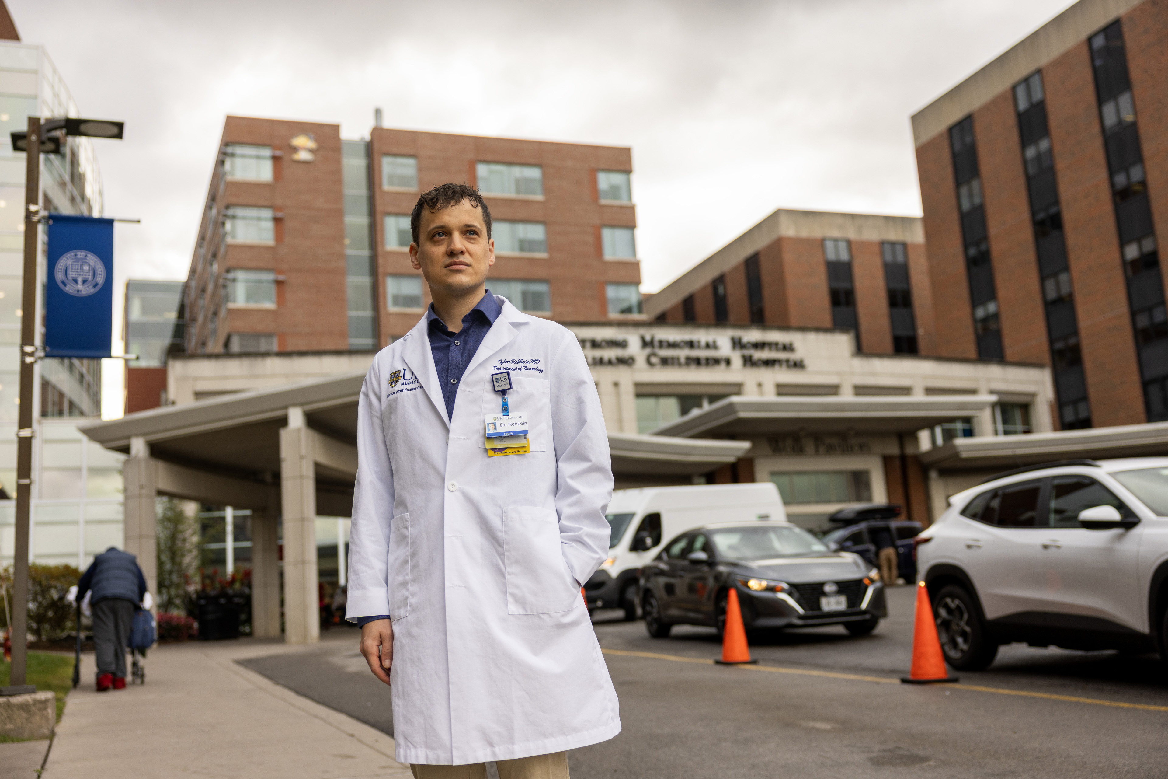 A physician wearing a white coat stands outside a hospital entrance