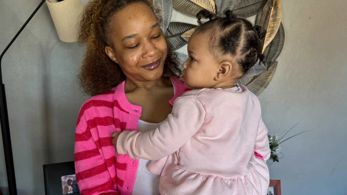 A Black woman holds her young daughter in her arms. She is smiling at her daughter.