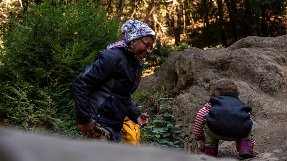 A senior woman in active outdoor clothing encourages a toddler to navigate uneven ground in a lush forest environment.