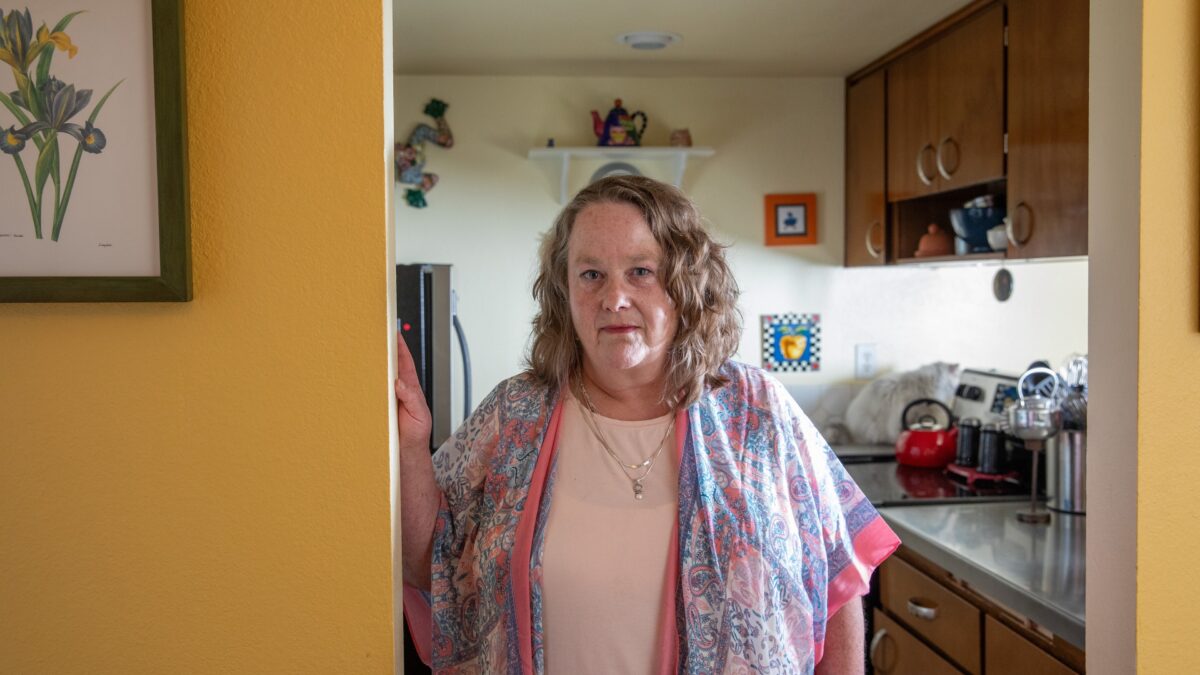A woman in a pink tshirt and a paisley pink and blue cardigan stands in the doorway of a kitchen beside a yellow wall
