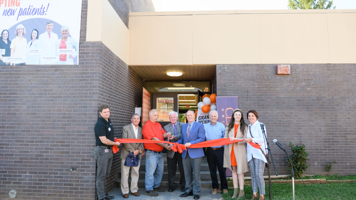 A photo showing a group of people standing in front of a brick building, behind a red ribbon, for an opening ceremony on a sunny day.