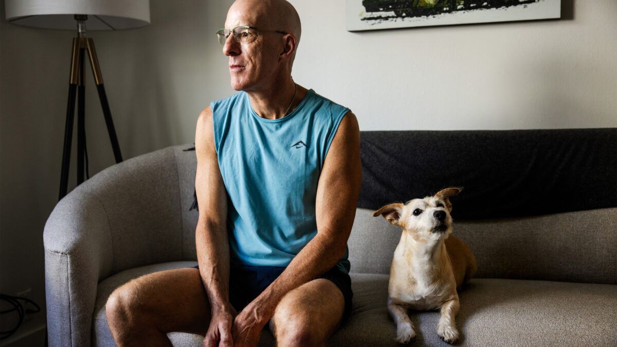 A man sits on a sofa next to a dog. Harsh light is cast on his face from a window out of frame.