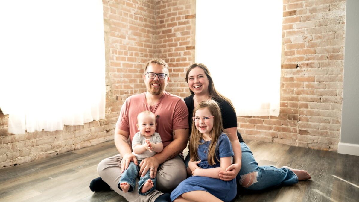 A mother and father sit on the floor in a well-lit room with their two young children in their laps for a studio portrait.