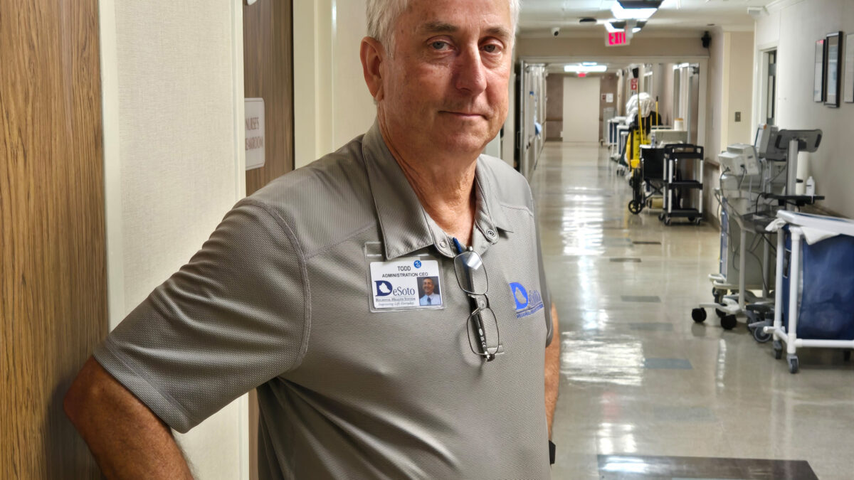 A photo of an older man standing for a portrait in a hospital corridor.