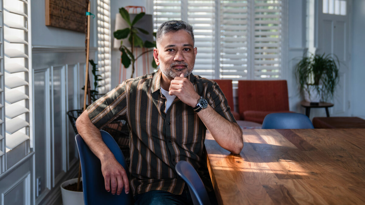 A South Asian man sits by the window at a table. He rests his elbow on the table with his hand on his chin. He looks contemplative.