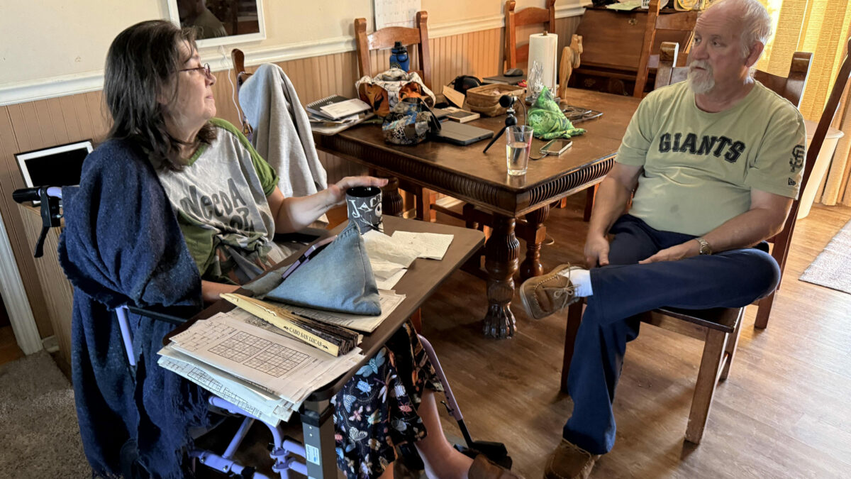 A husband sits in a chair conversing with his wife, who is in a wheelchair.