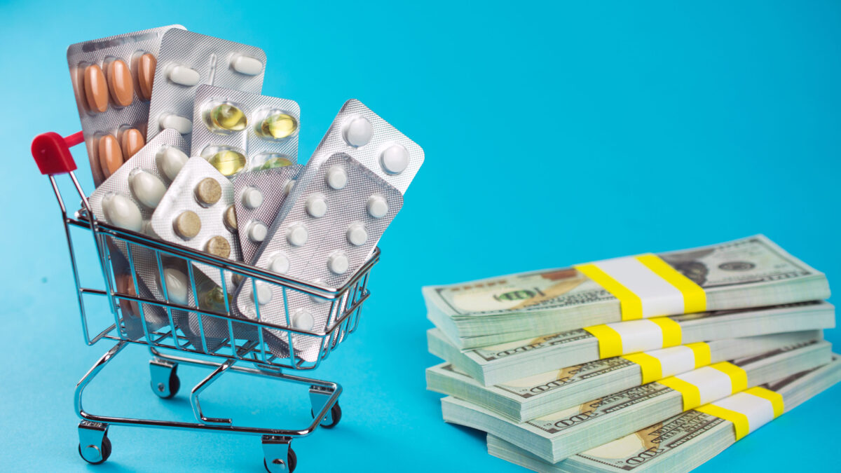A photo of a mini shopping cart filled with pills beside a stack of money against a blue background.